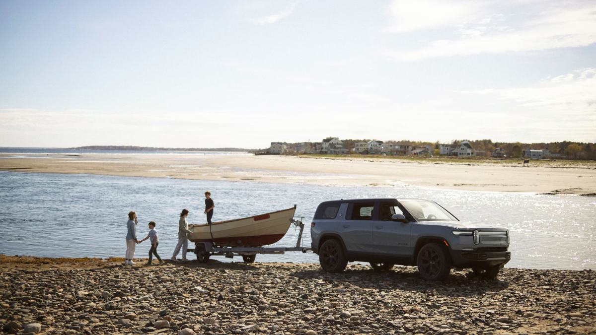 A silver Rivian R1T electric pickup truck is shown from the side view at a beach, backing a boat into the water while several people assist with the launch.